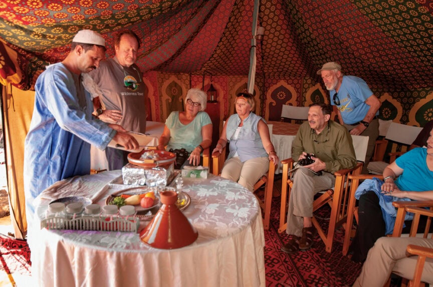 A man in traditional attire pours tea for a group of people seated around a table in a decorated tent. Various items, including tea sets and fruit, are arranged on the table, reflecting the rich traditions of world cuisine and delicious recipes from around the globe.