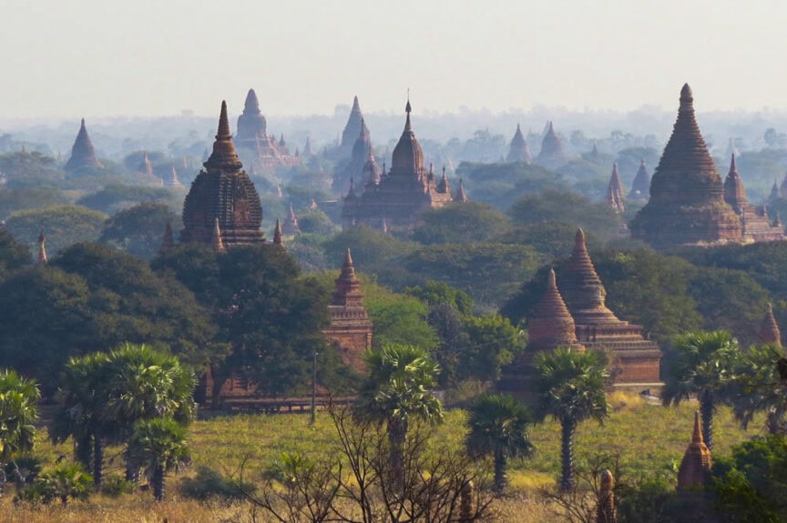 A landscape view of Bagan, Myanmar, showing the majestic ancient pagodas and temples surrounded by greenery and palm trees.