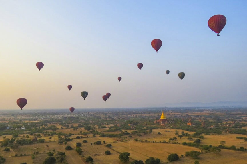 Hot air balloons float over the vast, mysterious landscape of Myanmar, with scattered vegetation and distant structures illuminated by the majesty of sunrise or sunset.