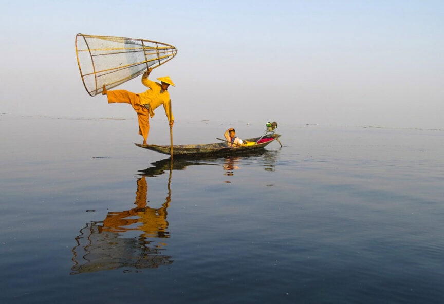 A person in a yellow outfit, emanating an air of mystery, balances on one leg on a canoe with a fishing net raised, while another individual sits serenely on the boat against the backdrop of Myanmar's majestic, calm waters.