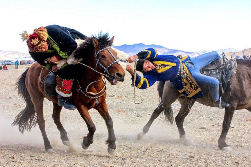 Two people in traditional attire participate in a horseback riding event during one of the amazing festivals around the world, both struggling to gain control while riding their horses on a rocky terrain.