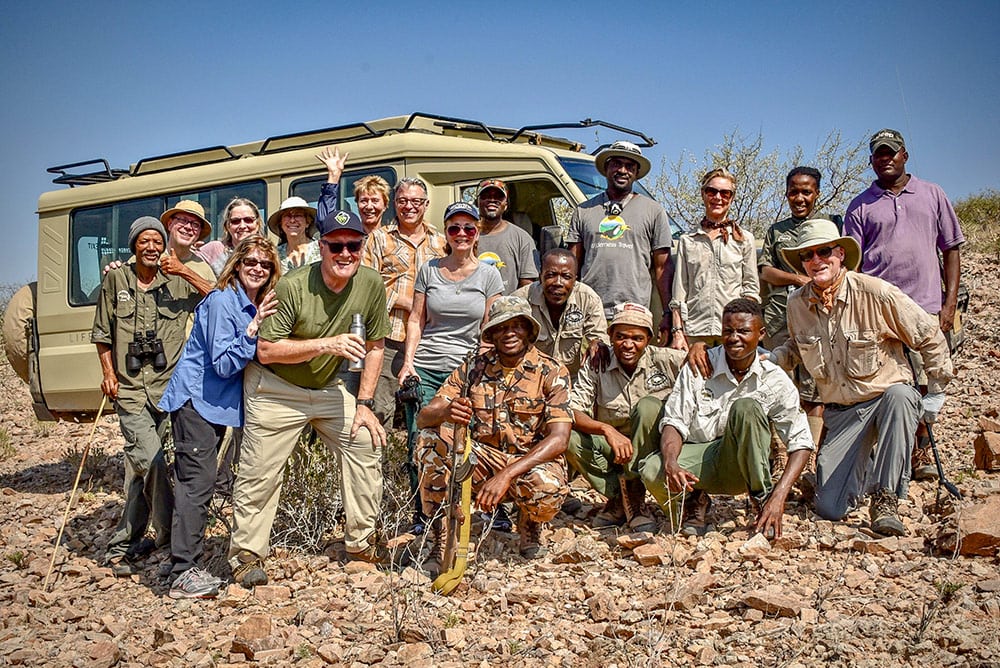 A group of 16 people, including tourists and wildlife conservationists, stand together in front of a safari vehicle on a rocky landscape in Namibia, smiling at the camera. Several hold walking sticks and binoculars, marking this special event with joy and camaraderie.