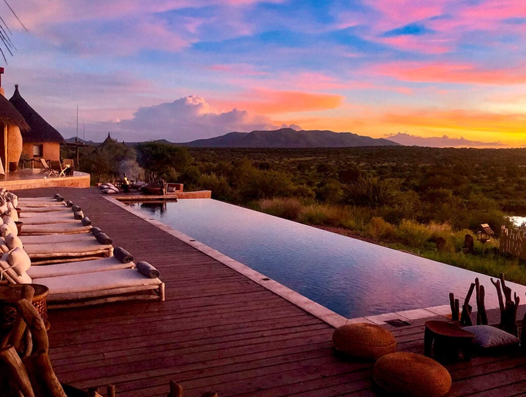A luxurious infinity pool on a wooden deck overlooks a lush green landscape and distant mountains under a vibrant sunset sky. Reserved lounge chairs and a fire pit are visible to the left, perfect for those exploring Namibia during the stunning Green Season.