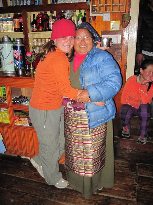 Two people pose happily for a photo in a cozy wooden room. One wears an orange hat and jacket, the other a blue jacket and traditional clothing from Nepal. A child in orange sits in the background, capturing a perfect moment for their family photobook.