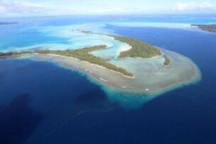 Aerial view of a tropical island in Palau surrounded by blue ocean waters and various shades of turquoise in a lagoon, featuring lush greenery and sandy beaches below.