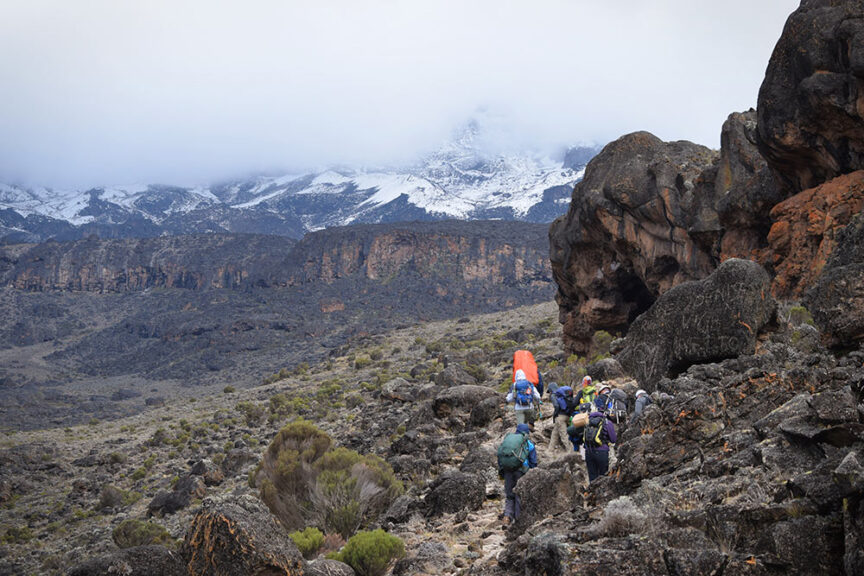A group of hikers with backpacks walk along a rocky trail in a mountainous area, reminiscent of those found when you hike Mt. Kilimanjaro in Tanzania, featuring snow-covered peaks and cloudy skies in the background.