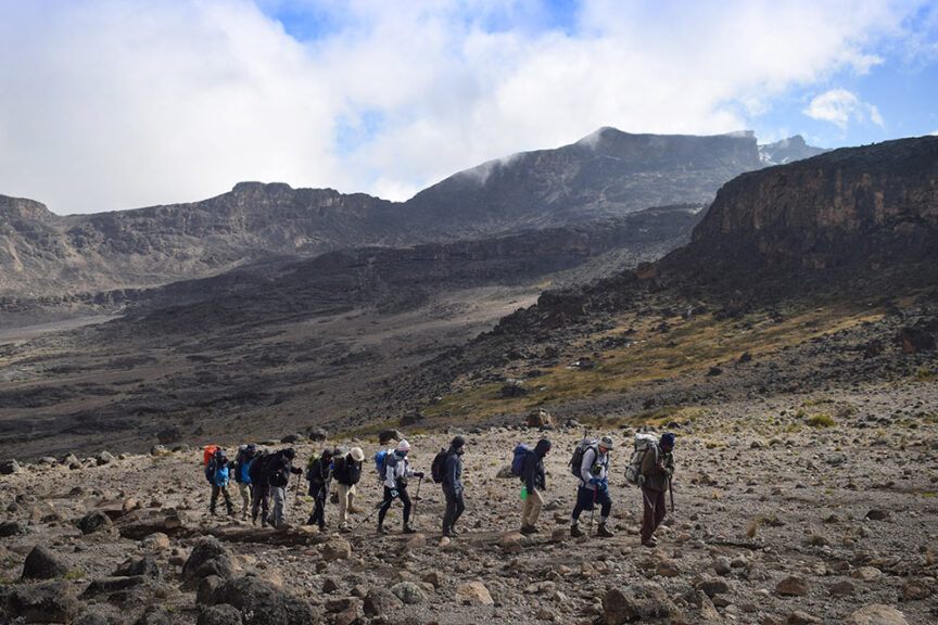 A group of people with backpacks hikes in a rugged, rocky landscape with distant mountains and cloudy sky overhead, reminiscent of the challenging trek to Hike Mt. Kilimanjaro in Tanzania.