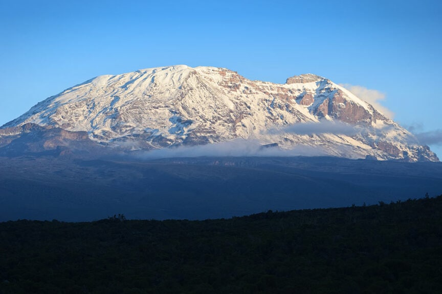 The image shows Mount Kilimanjaro with its snow-capped peaks and slopes, set against a clear blue sky and partially obscured by light clouds at the base. A dark green forest foreground in Tanzania is also visible, inviting adventurers to hike Mt. Kilimanjaro.