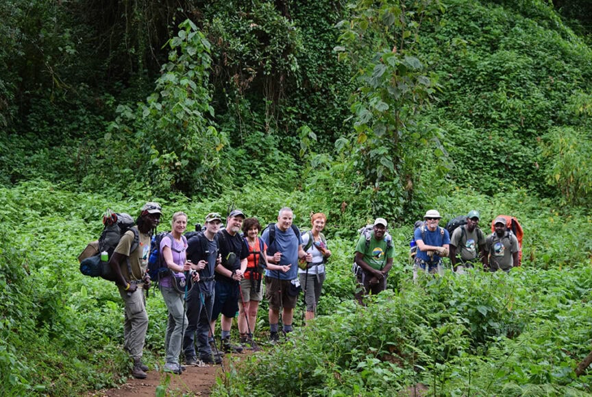 A group of people stands together on a forest trail in Tanzania, surrounded by lush green vegetation. Some are holding hiking poles and wearing backpacks, preparing for the best way to hike Mt. Kilimanjaro.