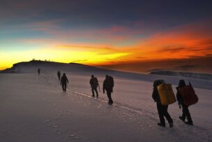 A group of hikers with backpacks trek up a snowy mountain at sunrise, leaving footprints in the snow, reminiscent of an adventurous hike up Mt. Kilimanjaro in Tanzania.