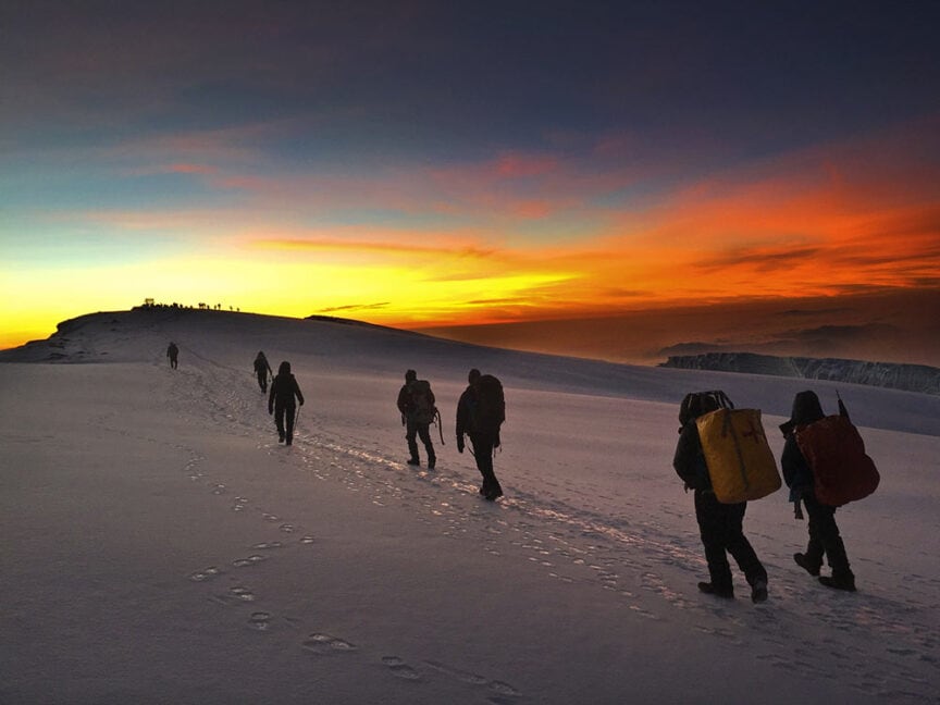 A group of hikers with backpacks trek up a snowy mountain at sunrise, leaving footprints in the snow, reminiscent of an adventurous hike up Mt. Kilimanjaro in Tanzania.