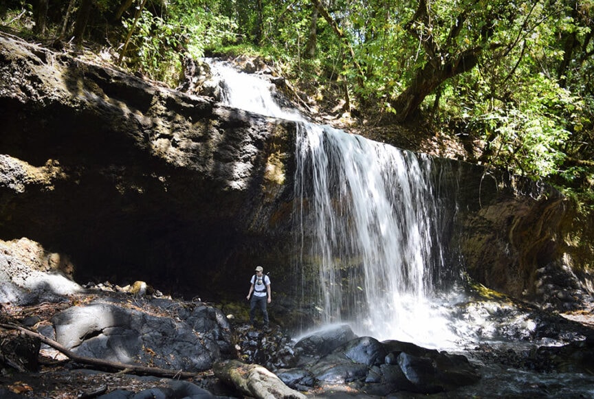 A person wearing a backpack stands on rocks near the base of a waterfall in a forested area, reminiscent of the lush trails you might encounter while hiking Mt. Kilimanjaro in Tanzania.
