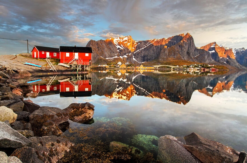 A red cabin stands on stilts by a calm lake with mountains in the background, reminiscent of European destinations. The scene is reflected in the water, showing the cabin, rocks, and surrounding landscape under a cloudy sky. A picturesque spot for any traveler type exploring Europe.