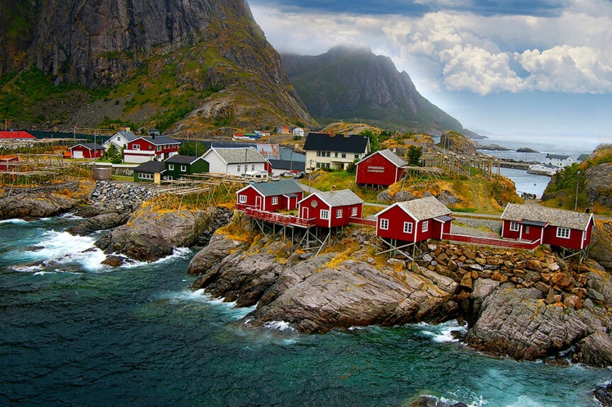 A coastal village with red and white houses on a rocky shoreline, surrounded by mountains and an overcast sky, near a body of water—a hidden gem for world travel enthusiasts seeking colorful cities.