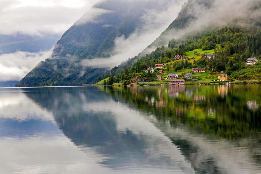 A serene lake reflects foggy mountains and scattered houses along a verdant shoreline under a cloudy sky, inviting travel adventures in one of the world's happiest countries.