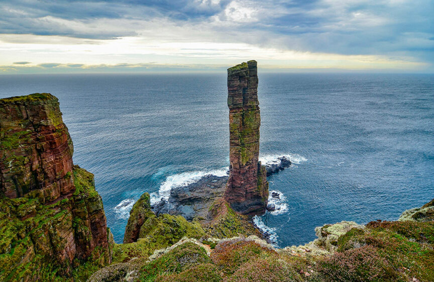 Tall sea stack in the ocean surrounded by rocky cliff edges and patches of green vegetation under a cloudy European sky, perfect for hiking enthusiasts looking to escape the crowds.