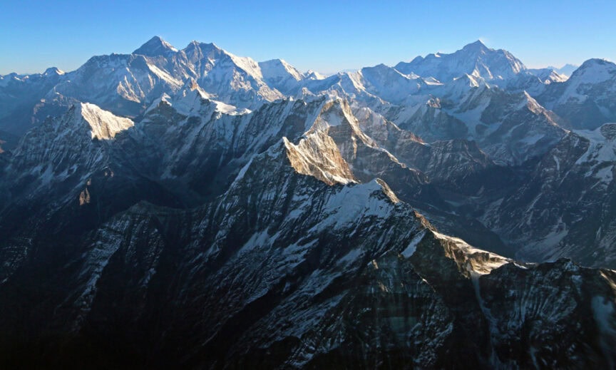 A panoramic bird's eye view of snow-capped mountain peaks under a clear blue sky, with sharp ridges and valleys visible. Captured through breathtaking aerial photography, the mountains are part of a wide-ranging, rugged landscape.