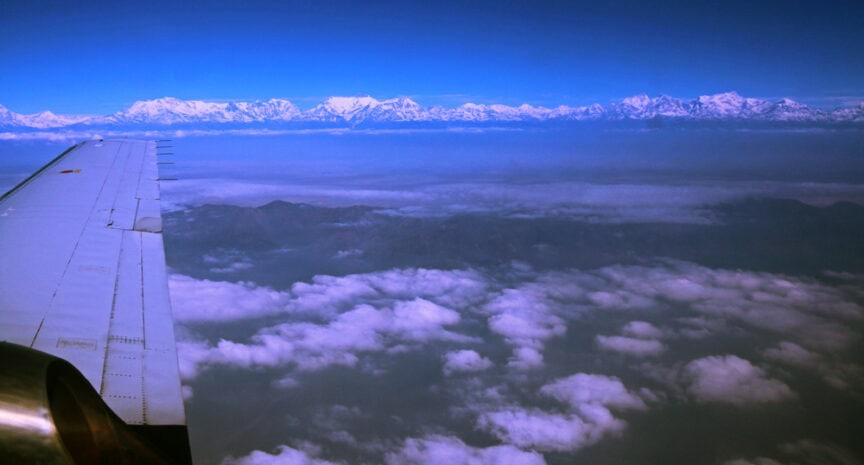 View from an airplane window showing a wing and engine with a vast expanse of clouds below and snow-capped mountain peaks on the horizon, offering a breathtaking bird's eye view of the majestic landscape.