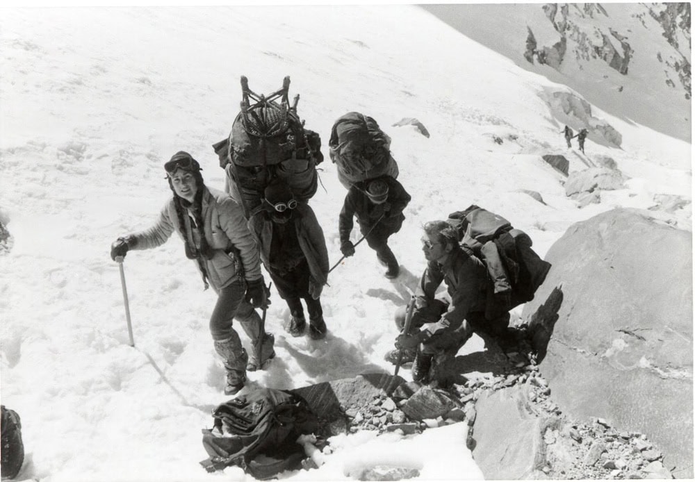 Three climbers, each carrying heavy packs, navigate a steep snowy mountain slope. One climber stands holding an ice axe, while the other two appear to adjust their gear. Other climbers are seen in the distance—a moment captured perfectly for the family photobook of their unforgettable expedition in Nepal.