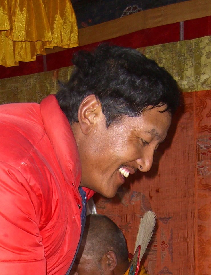 A person in a red jacket smiles while looking down, with a colorful background of drapes and a partially visible family member in the backdrop, capturing a joyful moment that would be perfect for a photobook of memories from Nepal.