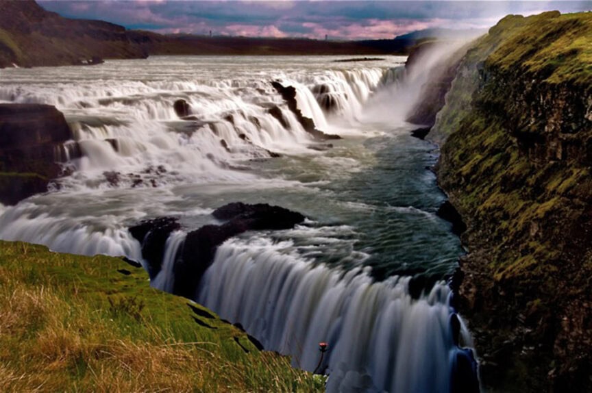 A powerful waterfall cascades over rocky cliffs into a river below, surrounded by the natural beauty of green hills under a cloudy sky.