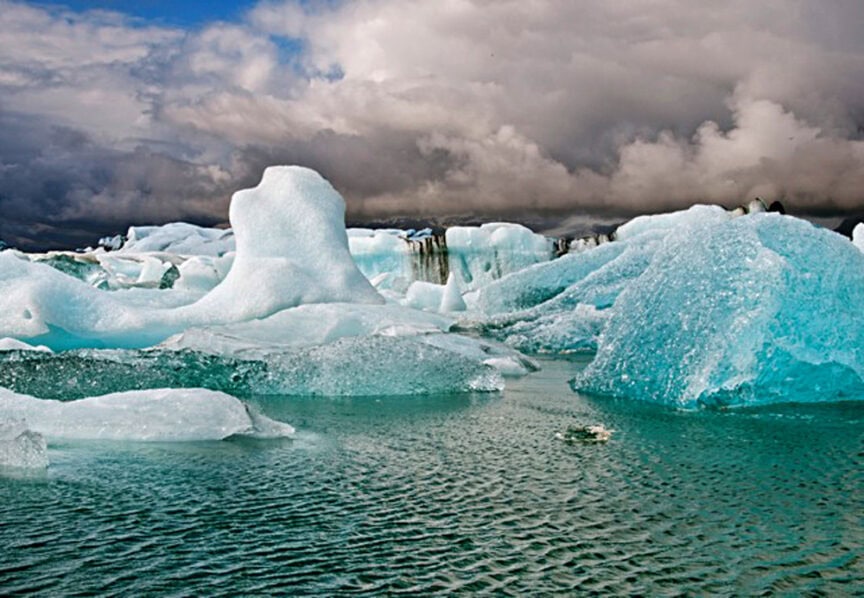 Icebergs float in a body of water under a cloudy sky, showcasing the natural beauty of their blue and white hues. They contrast dramatically with the dark, stormy clouds above.