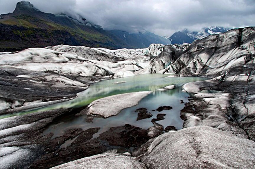 Glacial lagoon surrounded by ice and rocky terrain under cloudy skies, showcasing natural beauty with mountains in the background.