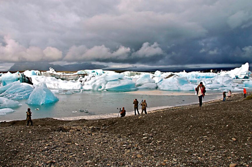 Several people stand and take photos, capturing the natural beauty of a lake surrounded by icebergs under a cloudy sky.