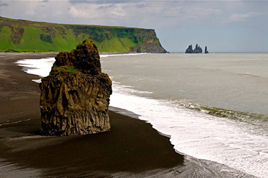 Rock formations stand along the black sand beach, showcasing natural beauty with green cliffs and ocean waves in the background under a cloudy sky.