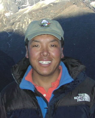 A smiling person wearing a beige cap and a blue jacket with "The North Face" logo stands outdoors in Nepal's majestic mountains, ready to capture moments for a family photobook.