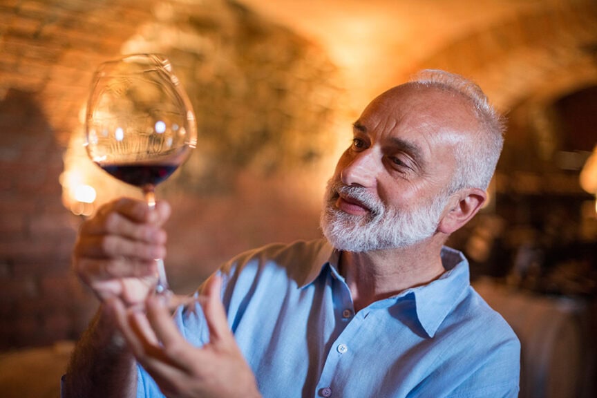 A man with a grey beard and blue shirt examines a glass of red wine in a warmly lit, rustic setting, reminiscing about his adventures in famous wine-growing regions.