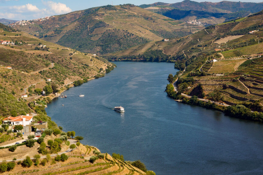 A river flows through a valley with terraced hillsides and a few boats on the water. Small clusters of houses can be seen along the riverbank, reminiscent of Portugal's scenic hiking trails, where Port Wine vineyards dot the landscape.