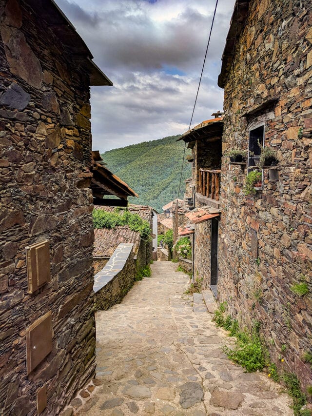 A narrow stone pathway runs between rustic stone buildings under a cloudy sky, leading towards a green hillside in the background, reminiscent of the scenic trails of Portugal. Perhaps after a day of hiking, one might indulge in a glass of Port Wine.