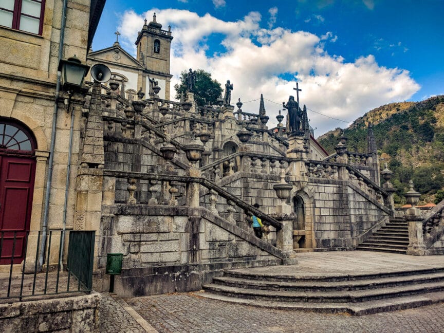 Ornate stone staircase with multiple levels and decorative railings, leading up to a historic building in Portugal against a backdrop of trees and a partly cloudy sky. After a phenomenal hiking adventure, enjoy some local port wine while soaking in the beauty of this picturesque scene.