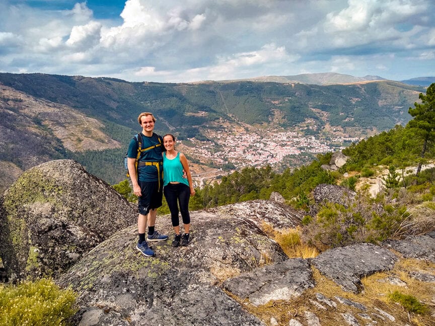 A man and woman in hiking attire stand on a large rock overlooking a valley with a town nestled among hills and trees under a partly cloudy sky, relishing the phenomenal hiking experience in Portugal.