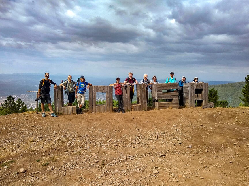 A group of people, after hiking a rugged trail, stands in front of large wooden letters spelling "URBASA" on a rocky hilltop, with a cloudy sky behind them. The scene evokes the adventurous spirit akin to exploring Portugal’s scenic terrains and savoring the richness of Port Wine.