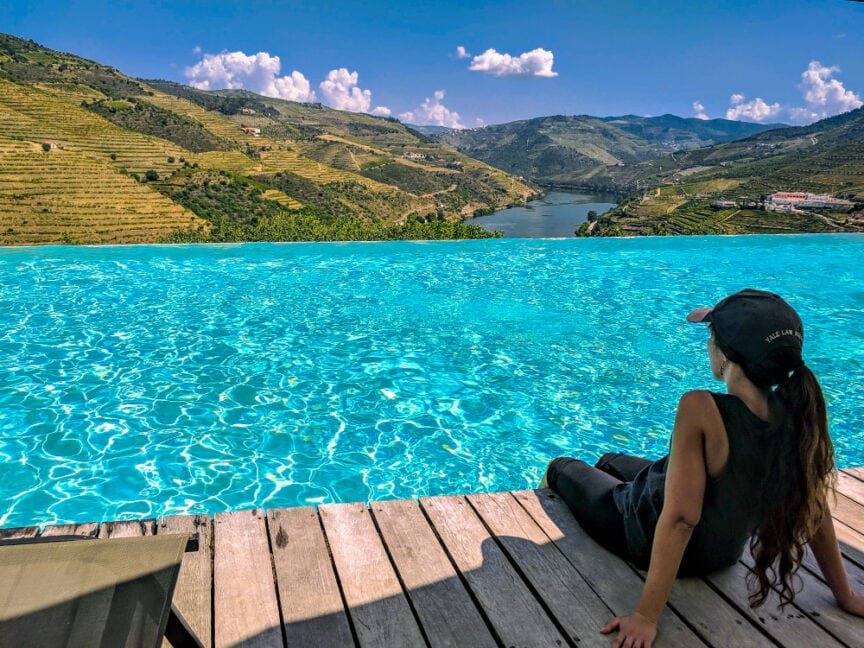 Person sits at the edge of an infinity pool in Portugal, overlooking a scenic valley with a river and terraced hills in the background under a partly cloudy sky, perhaps enjoying some Port Wine after a day of hiking.