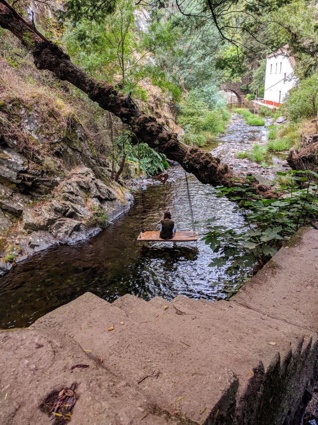 A person sits on a wooden swing suspended over a shallow creek surrounded by trees and rocky terrain, with a white building visible in the background—an idyllic spot reminiscent of Portugal's phenomenal hiking trails.