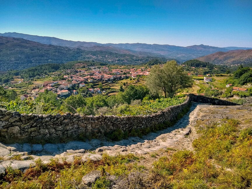 A stone pathway leads down a hill overlooking a village nestled between rolling green hills and mountains under a clear blue sky, reminiscent of the scenic trails in Portugal. This picturesque route is perfect for hiking enthusiasts, offering breathtaking views that pair as perfectly as port wine with local delights.