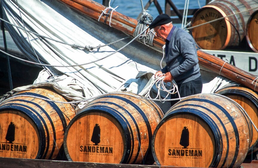 A man in a dark cap and jacket ties ropes on large wooden barrels labeled "Sandeman" on a docked sailboat, preparing for an adventure through wine-growing regions.