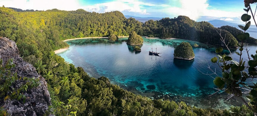 A secluded bay with clear turquoise water surrounded by lush green hills in Raja Ampat. A boat is anchored near small rocky islets in the middle of the bay, inviting explorers to embark on underwater adventures.
