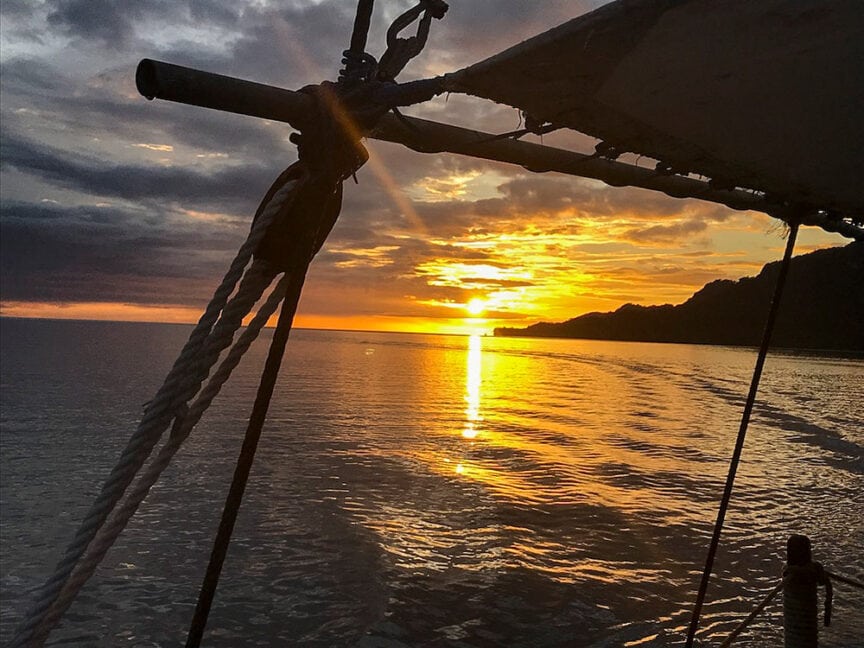 A boat sail and rigging frame a vibrant sunset over the calm sea waters of Raja Ampat, with the silhouette of a coastal landscape in the background, hinting at rich underwater adventures waiting to be explored.
