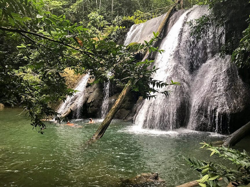 A waterfall cascades into a green pool surrounded by lush foliage, with people swimming near the base of the falls, reminiscent of the tranquil scenes in Raja Ampat's underwater adventures.