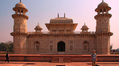 A person stands in front of the Tomb of I'timād-ud-Daulah in Agra, India, capturing the intricate white marble facade and minaret-style towers on video. Another person is visible walking on the left, adding to this family's adventure in vibrant India.