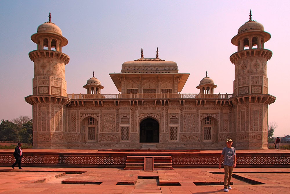 A person stands in front of the Tomb of I'timād-ud-Daulah in Agra, India, capturing the intricate white marble facade and minaret-style towers on video. Another person is visible walking on the left, adding to this family's adventure in vibrant India.