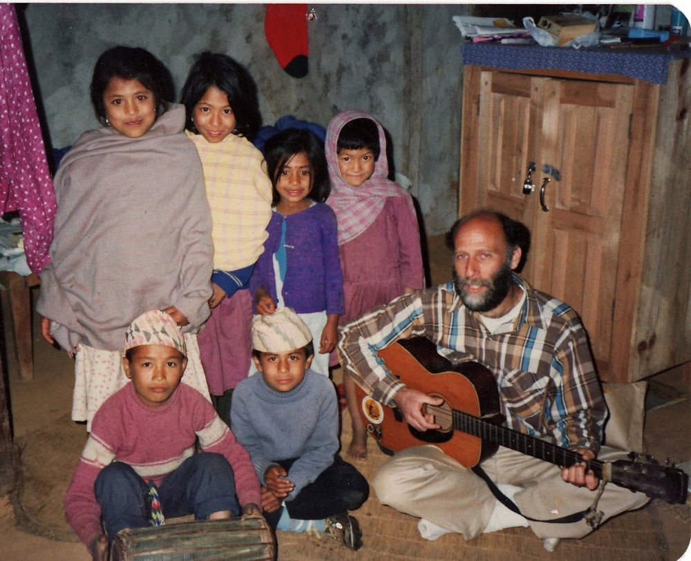 A group of six children poses indoors beside a man playing a guitar, creating a heartwarming photobook moment. The room, reminiscent of family gatherings in Nepal, features a wooden cabinet and various items around. The children are dressed in casual, warm clothing.