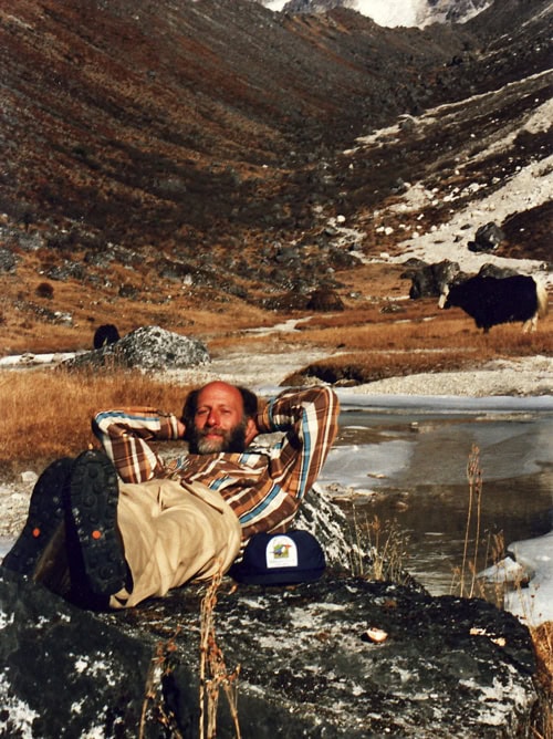 A man in a plaid shirt and khaki pants lounges on a rock in a mountainous, grassy area with a river and hills in the background, creating the perfect scene for a family photobook reminiscent of adventures in Nepal.