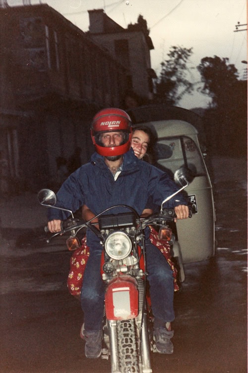 Two individuals ride a motorcycle with the driver wearing a red helmet and the passenger smiling, seen on a street in Nepal with buildings and a vehicle in the background—an ideal moment for any family photobook.