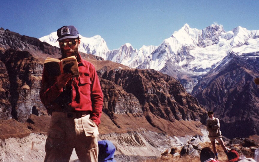 A person in a red shirt and sunglasses stands reading a book in front of a mountainous landscape with snow-capped peaks, reminiscent of a perfect family photobook shot. Another person and camping gear are visible in the background, capturing memories likely from an adventurous trip in Nepal.