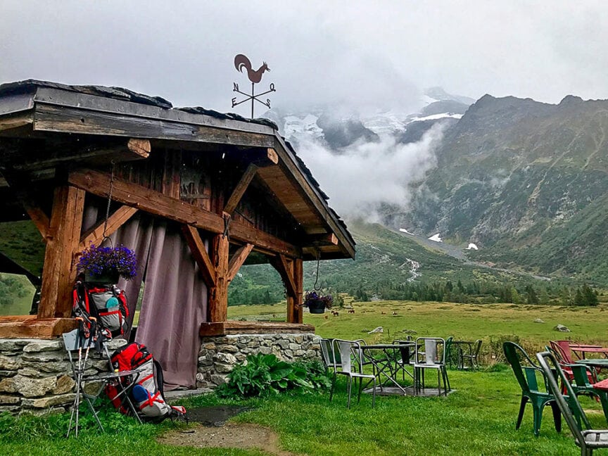 A rustic wooden cabin with backpacks resting against it stands in front of mountainous terrain and misty clouds, evoking the spirit of hiking the Tour du Mont Blanc. Green chairs and tables are arranged on the grass nearby, inviting weary travelers to rest and enjoy the serene scenery.
